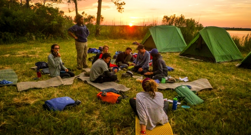 A group of young people rest on pads in the grass near their tents. The sun is setting behind a body of water in the background. 